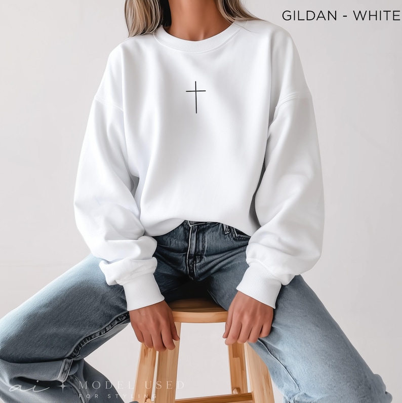 Puede incluir: Una sudadera blanca de cuello redondo marca Gildan con una peque&ntilde;a cruz negra centrada en el pecho. El su&eacute;ter de manga larga tiene cuello, pu&ntilde;os y dobladillo acanalados, combinado con jeans de mezclilla azul claro. El texto GILDAN - WHITE es visible.