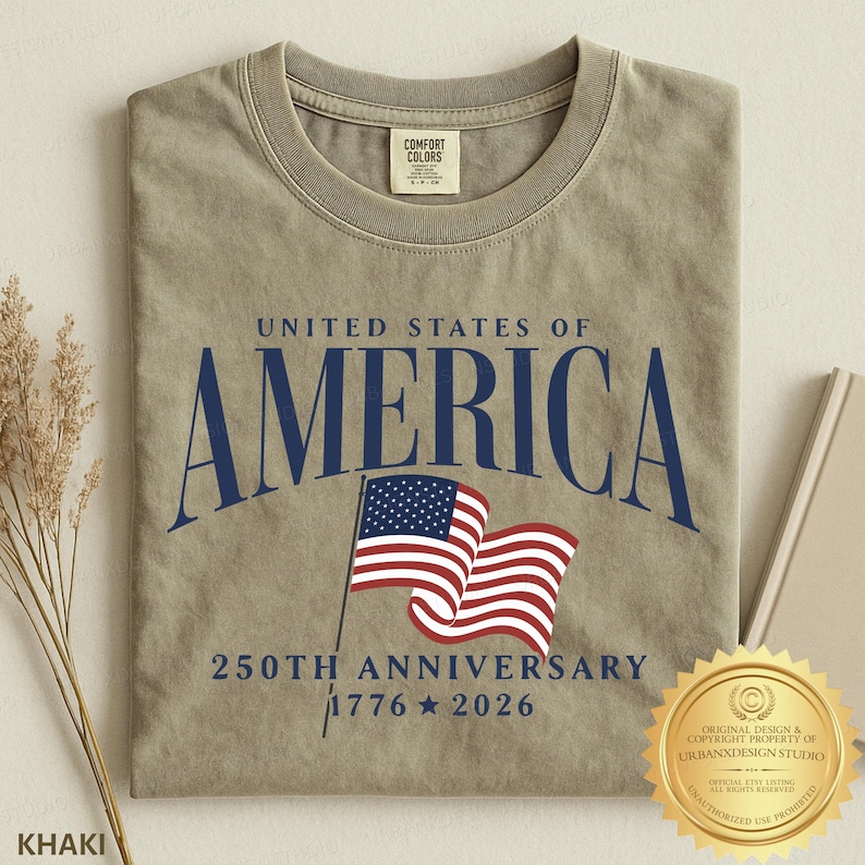 May include: A khaki-colored crew neck t-shirt featuring the dark blue text UNITED STATES OF AMERICA, a graphic of a waving American flag, and the text 250TH ANNIVERSARY 1776 2026. The shirt has a Comfort Colors brand label on the inner neck collar.