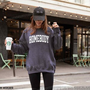 May include: A dark heather grey sweatshirt with the word "HOMEBODY" printed in white on the front. The sweatshirt is being worn by a person holding a to-go coffee cup.