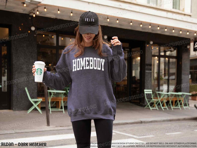 May include: A dark heather grey sweatshirt with the word "HOMEBODY" printed in white on the front. The sweatshirt is being worn by a person holding a to-go coffee cup.