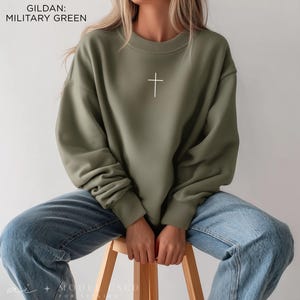 Puede incluir: Una sudadera de cuello redondo Gildan en color verde militar con un peque&ntilde;o gr&aacute;fico de una cruz blanca centrado en el pecho. El jersey de manga larga tiene cuello, pu&ntilde;os y dobladillo acanalados, combinado con pantalones vaqueros azul claro.