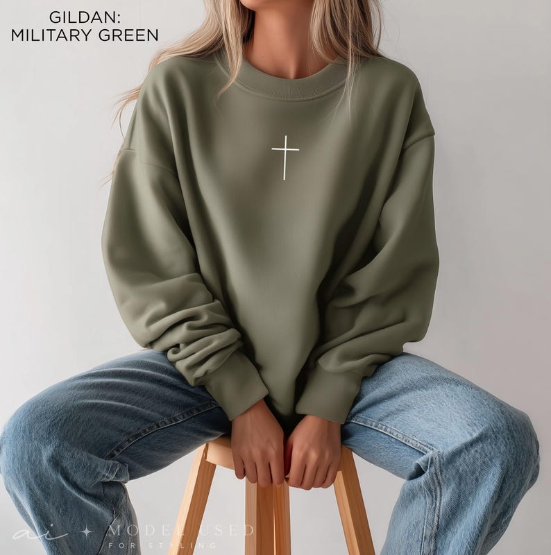 Puede incluir: Una sudadera de cuello redondo Gildan en color verde militar con un peque&ntilde;o gr&aacute;fico de una cruz blanca centrado en el pecho. El jersey de manga larga tiene cuello, pu&ntilde;os y dobladillo acanalados, combinado con pantalones vaqueros azul claro.