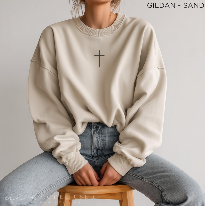 Puede incluir: Una sudadera de cuello redondo color arena con un peque&ntilde;o gr&aacute;fico de una cruz negra centrado en el pecho. La prenda tiene mangas largas con pu&ntilde;os acanalados y un cuello acanalado. El texto GILDAN - SAND est&aacute; impreso en la esquina superior derecha.