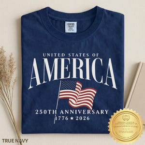 May include: A navy blue t-shirt featuring the white text UNITED STATES OF AMERICA, a graphic of a waving American flag, and the text 250TH ANNIVERSARY 1776 2026. The shirt has a Comfort Colors brand label on the inside of the crew neckline.