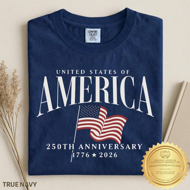 May include: A navy blue t-shirt featuring the white text UNITED STATES OF AMERICA, a graphic of a waving American flag, and the text 250TH ANNIVERSARY 1776 2026. The shirt has a Comfort Colors brand label on the inside of the crew neckline.