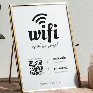 Puede incluir: Un letrero enmarcado con un marco de metal dorado. El letrero tiene las palabras "wifi is on the house!" en negro, junto con un código QR e información de la red. Un pequeño frasco de vidrio y un jarrón con vegetación están en el fondo.