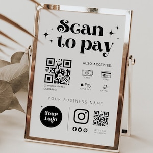 Puede incluir: Un cartel blanco con texto y gráficos negros, que dice "Scan to pay". El cartel también incluye un código QR, un logotipo e iconos para efectivo, tarjeta, Apple Pay y PayPal. El cartel está en un marco plateado y está sobre una superficie blanca.