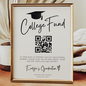 May include: A framed sign with the words "College Fund" and a QR code. The sign is in a gold frame and sits on a wooden table. The sign also includes the text "Evelyn's Graduation" and a graduation cap graphic.