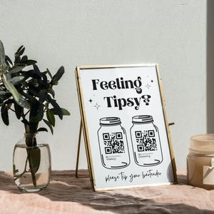 May include: A white sign with gold trim and a black and white illustration of two mason jars with QR codes. The sign reads "Feeling Tipsy? Please tip your bartender."