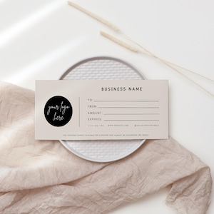 Op de afbeelding: Een beige cadeaubon met zwarte tekst. De tekst luidt "BUSINESS NAME", "TO", "FROM", "AMOUNT", "EXPIRES", "WWW.WEBSITE.COM" en "@SOCIALMEDIANHANDLE". Een zwarte cirkel met de tekst "your logo here" bevindt zich in de linkerbovenhoek.