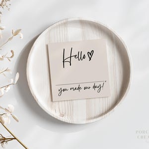 Op de afbeelding: Een witte kaart met zwarte kalligrafie met de tekst "Hello &hearts; you made our day!" De kaart ligt op een wit houten bord.