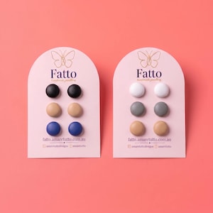 Puede incluir: Dos tarjetas de presentación arqueadas con la etiqueta Fatto handmade jewellery, cada una con tres pares de pendientes circulares. La tarjeta izquierda tiene pendientes negros, beige y azules. La derecha tiene pendientes blancos, grises y beige. Incluye fatto.amaretutto.com.au.