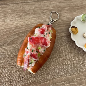 May include: A lobster roll keychain made of soft, squishy material. The keychain is shaped like a lobster roll with a white bun, red lobster meat, and green chives. The keychain has a silver metal ring.
