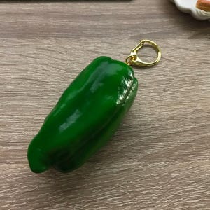 May include: A green bell pepper-shaped keychain with a gold-colored clasp. The keychain is made of a glossy, solid green material. The background is a wooden surface.