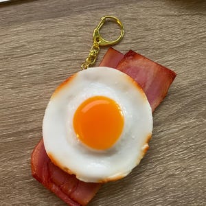 May include: A novelty keychain featuring a fried egg with a bright orange yolk and white egg whites, atop two strips of red bacon. The keychain has a gold-colored metal ring and chain.