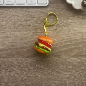 May include: A miniature burger keychain with a gold-colored ring. The burger features a sesame seed bun, a tomato slice, cheese, lettuce, and a patty. The keychain is on a wooden surface.