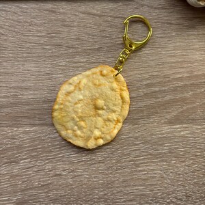May include: A unique keychain featuring a circular, textured charm resembling a fried food item. The charm is a light golden-brown color and is attached to a gold-colored keyring and chain. The background includes a keyboard and a wooden surface.
