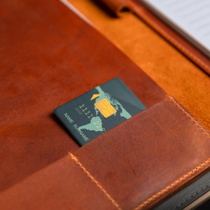 Pu&ograve; includere: Una cartella in pelle marrone con una carta di credito infilata nella tasca. La carta ha un design a mappa del mondo e il testo "NAME SURNAME" e "1111 1233 22".