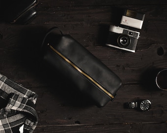 May include: A black leather zippered pouch with a gold zipper, a black and white plaid shirt, a black camera with a flash, a black watch, and a black cup on a dark wood surface.
