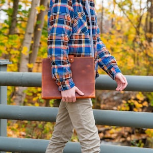 Pu&ograve; includere: Una persona che indossa una camicia a quadri blu e rossi e pantaloni kaki tiene in mano una cartella in pelle marrone. La persona sta camminando su un ponte con una ringhiera di metallo.