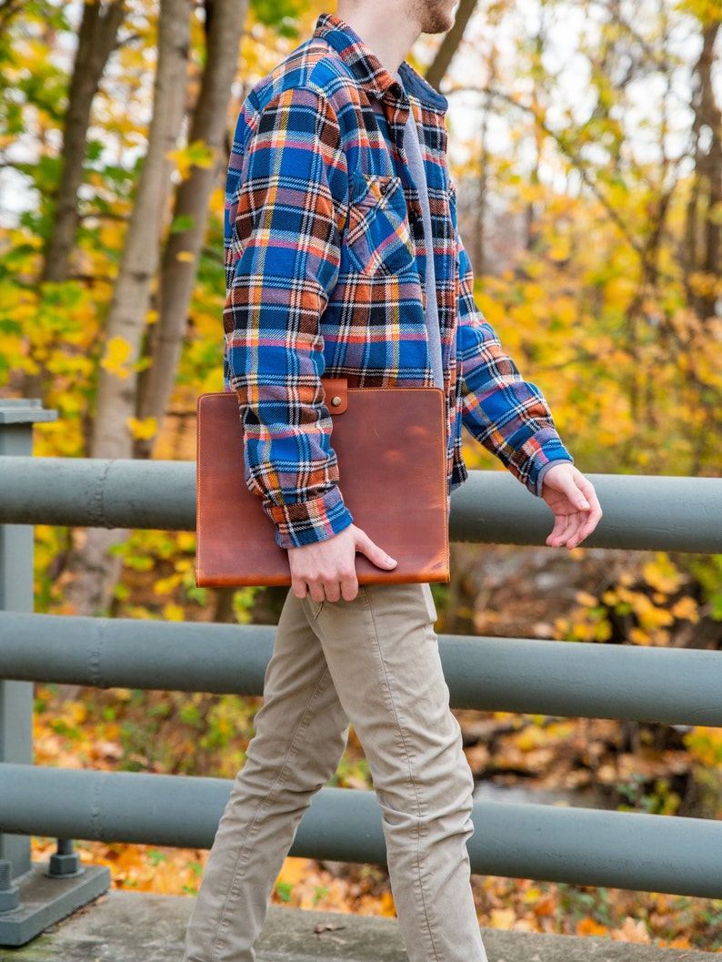 Pu&ograve; includere: Una persona che indossa una camicia a quadri blu e rossi e pantaloni kaki tiene in mano una cartella in pelle marrone. La persona sta camminando su un ponte con una ringhiera di metallo.