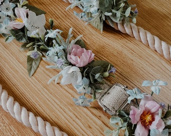 May include: Close-up of floral dog collar with a cream-colored rope design. The collar features pink, white, and blue flowers with green leaves. A silver-colored buckle is visible. The background is a light brown wooden surface.