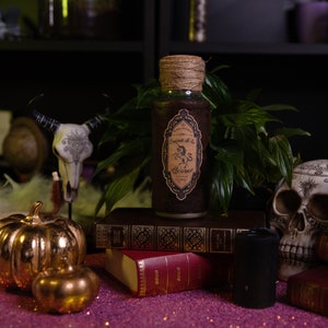 Pode incluir: Uma garrafa de vidro com um rótulo castanho que diz "Elixir of the Unicorn" e "Behemoth". A garrafa está sentada em cima de uma pilha de livros com uma caveira e uma abóbora dourada em primeiro plano.