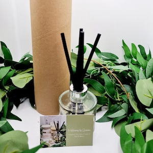 May include: A clear glass essential oil diffuser with black reeds and a silver collar. A brown cardboard tube stands behind the diffuser. A business card and green foliage are in the foreground. The card reads "Melloway by Gemma".