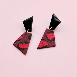 Peut inclure: Une paire de boucles d'oreilles géométriques. Chaque boucle d'oreille présente un dessus triangulaire noir et un bas triangulaire bordeaux avec un motif rouge et noir. Les boucles d'oreilles sont reliées par une petite boucle dorée.