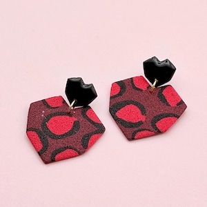 Peut inclure: Une paire de boucles d'oreilles géométriques. Les boucles d'oreilles présentent un dessus noir en forme de lèvres et un hexagone bordeaux avec un motif tacheté rouge et noir. Elles sont suspendues à un crochet doré sur fond rose.