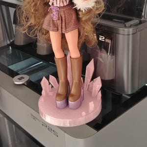 May include: A fashion doll with long brown hair, wearing orange-tinted glasses, a pink and brown striped long-sleeve top with white faux fur cuffs and collar, a brown skirt, and brown platform boots, standing on a circular pink crystal-like base.