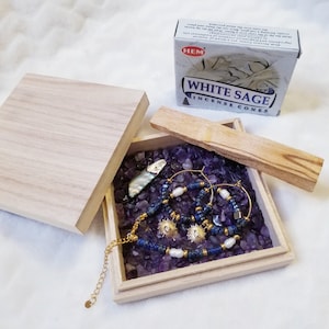 Peut inclure: Une boîte en bois avec un couvercle ouvert, révélant une petite coquille d'ormeau, deux créoles en or, un bracelet en chaîne dorée avec des perles bleues et blanches, et un petit sachet de cônes d'encens à la sauge blanche. La boîte est remplie de cristaux d'améthyste.
