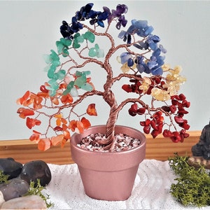 Peut inclure: Un arbre en cristal arc-en-ciel avec une base en fil de cuivre et un pot rose. L'arbre est entouré de sable blanc et de petites pierres grises. Une petite statue de Bouddha noire se trouve à droite de l'image.