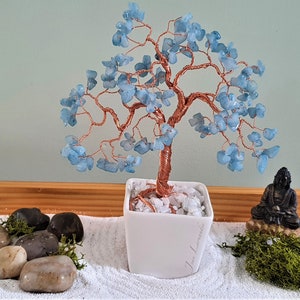 May include: A decorative bonsai tree with a copper wire trunk and branches, adorned with light blue gemstone leaves. The tree sits in a white square pot filled with white stones. A small Buddha statue and stones are nearby.