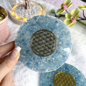May include: Two round blue crystal coasters with a gold flower of life design in the center. The coasters are made of blue aquamarine crystals and are set on a white fluffy surface.