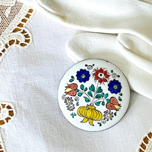 May include: A round, white enamel brooch with a floral design. The brooch features a yellow vase with a bouquet of red, blue, and orange flowers. The brooch is set against a white background.