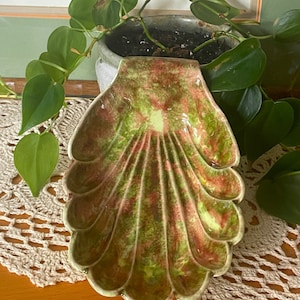 May include: A green and pink ceramic shell-shaped dish with a scalloped edge. The dish is glazed with a mottled finish and has a glossy sheen.