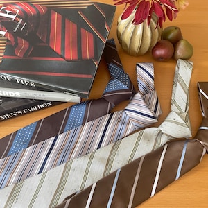 Puede incluir: Una variedad de corbatas con diferentes patrones y colores, incluyendo rayas y lunares, se exhiben sobre una superficie de madera. También se ven libros sobre corbatas y moda masculina, junto con un jarrón decorativo y fruta.