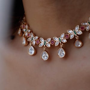 Puede incluir: Un collar gargantilla de oro con un diseño floral con piedras preciosas blancas, verdes y rojas. El collar tiene colgantes en forma de lágrima con piedras transparentes.