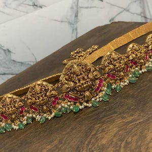 May include: Gold-coloured belt with intricate, detailed designs. The belt features ornate, sculpted figures with red and green accents. Small green beads and white pearls are strung along the bottom edge of the belt, adding a touch of elegance. The belt is displayed on a wooden surface.