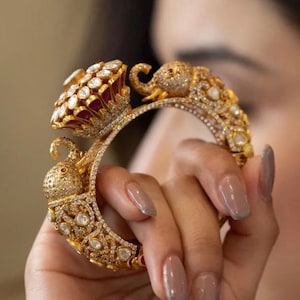 May include: A gold bangle bracelet with intricate detailing, featuring two elephant figures and a central lotus flower design. The bracelet is adorned with numerous sparkling white gemstones.