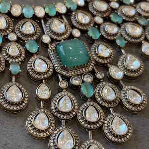 May include: A silver and green statement necklace with a large emerald green stone in the centre. The necklace is made of multiple layers of small round stones and teardrop-shaped stones.