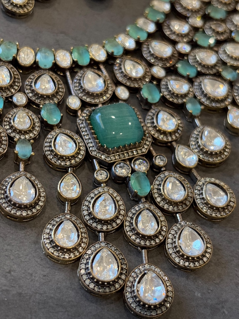 May include: A silver and green statement necklace with a large emerald green stone in the centre. The necklace is made of multiple layers of small round stones and teardrop-shaped stones.