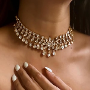 Puede incluir: Un collar gargantilla de oro con un gran colgante adornado en forma de lágrima. El colgante está hecho de piedras transparentes y está rodeado de piedras más pequeñas. El collar es una hermosa pieza de joyería que sería perfecta para una ocasión especial.