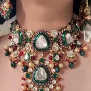 May include: A traditional Indian necklace with a large, white gemstone surrounded by green and red stones. The necklace is made of gold and has a delicate, intricate design.