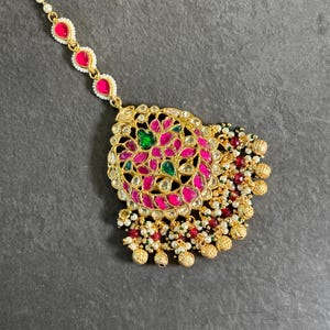 Puede incluir: Un maang tikka dorado con una pieza central redonda adornada con piedras rosas y verdes. El tikka tiene una cadena de perlas blancas y piedras rosas que conducen a la pieza central. La pieza central está rodeada por una franja de perlas blancas, piedras rosas y cuentas doradas.