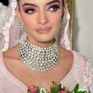 May include: A woman wearing a pink and white embroidered dress with a diamond necklace and earrings. She is holding a bouquet of pink and white roses.