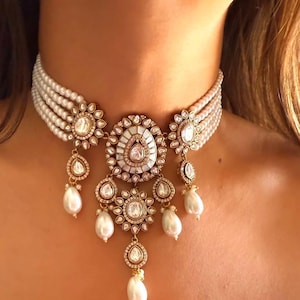 Puede incluir: Un collar gargantilla de perlas blancas con un colgante de oro que presenta un diseño circular grande y ornamentado con colgantes más pequeños en forma de pera. El colgante está decorado con piedras blancas y tiene una gota de perla blanca.