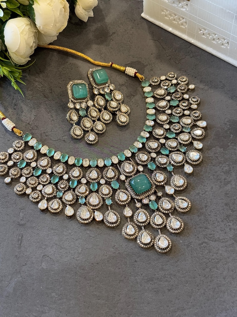 May include: A silver and green statement necklace with a large emerald green stone in the centre. The necklace is adorned with smaller emerald green stones and clear rhinestones. Matching earrings with a smaller emerald green stone are also included.