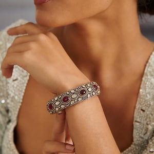 Op de afbeelding: Een gedetailleerde armband met een goudkleurige metalen basis, met afwisselend ovale en ronde rode edelstenen, en kleinere witte edelstenen. De armband wordt om een pols gedragen, wat het ingewikkelde ontwerp laat zien.